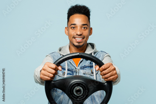 Young smiling happy fun cheerful man of African American ethnicity wear denim jacket orange t-shirt hold steering wheel driving car isolated on plain pastel light blue cyan background studio portrait.