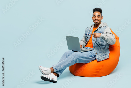 Full body young IT man of African American ethnicity wear denim jacket orange t-shirt sit in bag chair hold use work point finger on laptop pc computer isolated on plain pastel light blue background.