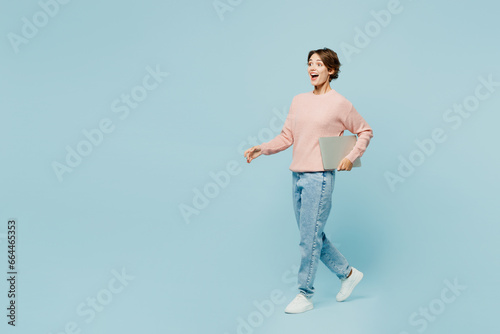 Full body side view young surprised IT woman she wear beige knitted sweater casual clothes hold use work on laptop pc computer isolated on plain pastel light blue cyan background. Lifestyle concept.