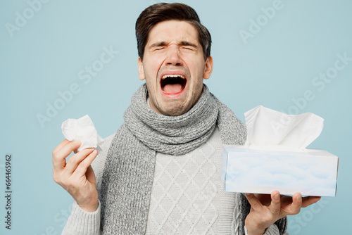 Young sad ill sick man wear gray sweater scarf sneezing hold box of paper napkins cry sneeze isolated on plain blue background studio. Healthy lifestyle disease treatment cold season recovery concept. photo