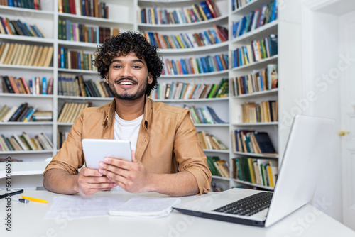 Photography Close-up photo of a young Indian male student working in a university library, sitting at a desk, using a tablet and smiling at the camera