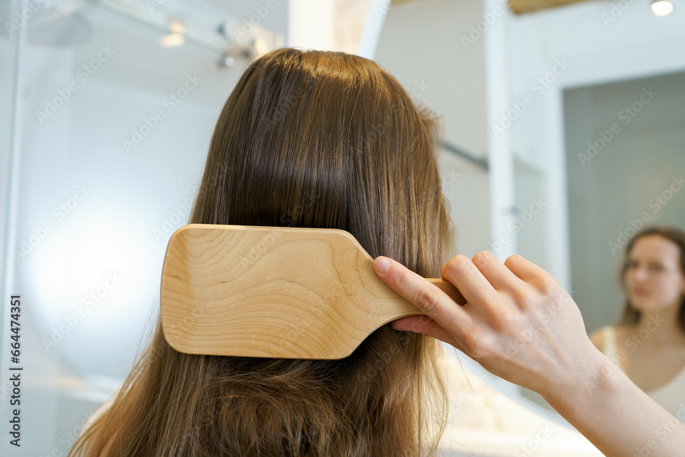 Fototapeta premium Close-up of a young brown-haired woman combing her hair with a wooden comb in the bathroom, rear view. The concept of hair cosmetics and products for shine