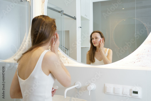 A young woman applies moisturizer to her face while looking in the bathroom mirror. Concept of beauty products for skin care, face cream