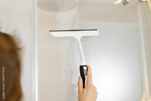 Close-up of a young woman using a scraper to remove drops from a glass shower and lime scale. The concept of cleaning in the apartment and in the bathroom photo