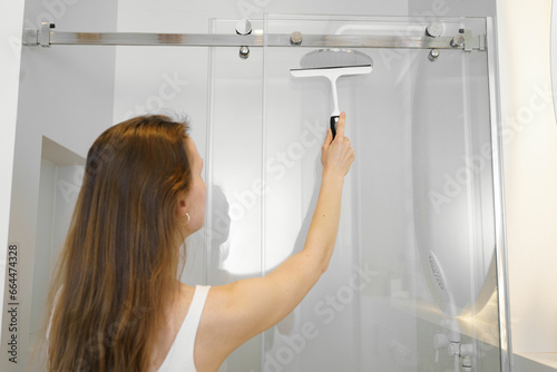A young woman cleans a glass shower cabin from splashes of water to remove limescale. Bathroom cleaning concept