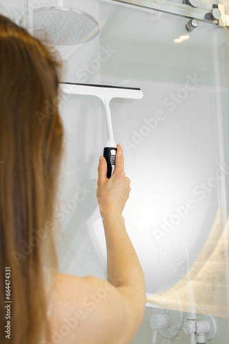 Vertical image of a young woman wiping the glass in the shower with a scraper from water drops to remove limescale. The concept of tidying up the apartment