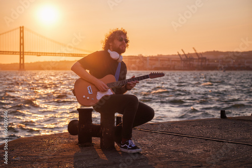 Hipster street musician in black playing electric guitar in the street on sunset on embankment with 25th of April bridge in background. Lisbon, Portugal