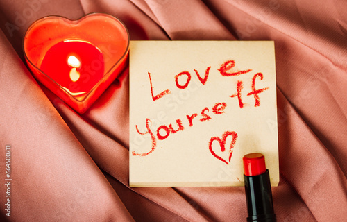 A note to self with a call to love yourself drawing with red lipstick on a silk terracotta cloth top view. A burning heart-shaped red candle, interior decor for women's home. Happy Valentines Day 2024