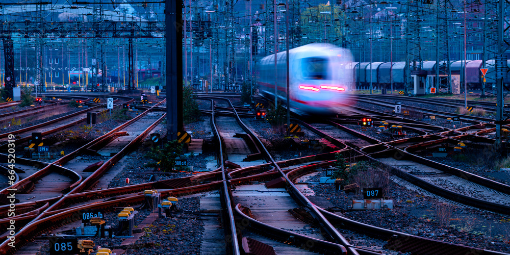 Railway panorama with leaving train in early morning twilight at blue hour. Tracks, oververhead lines, switches at Hagen main station in Ruhr basin, Germany. Transportation technology for passengers. - obrazy, fototapety, plakaty 