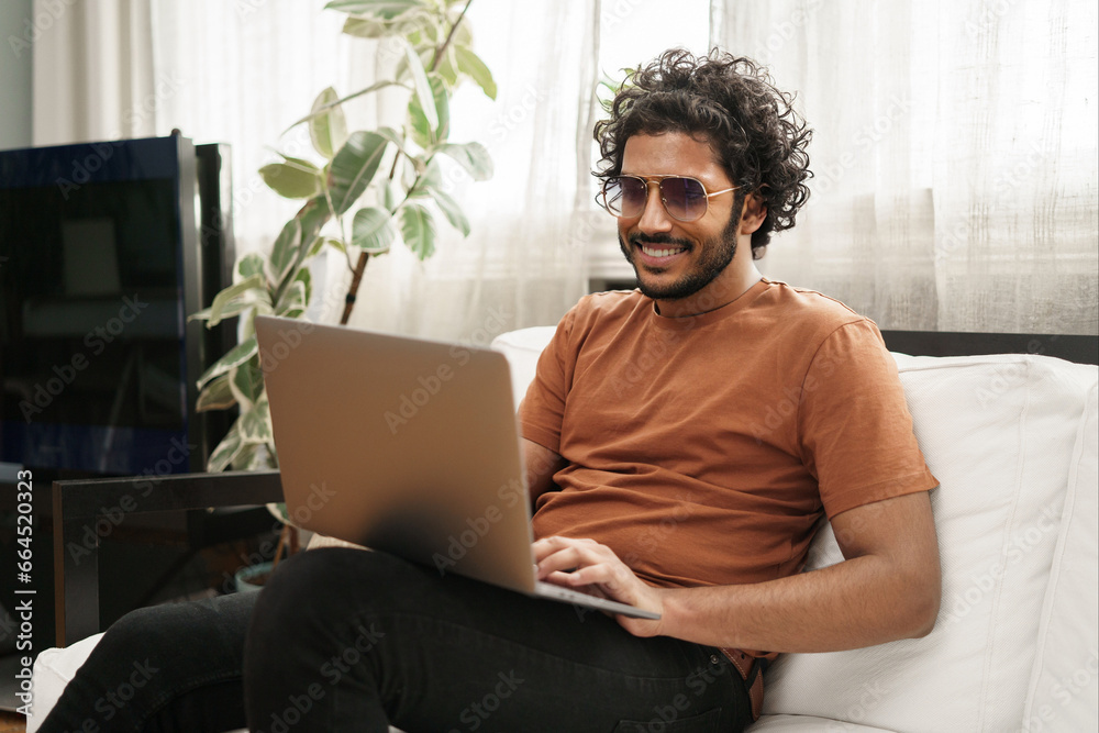 Happy male student working on a laptop while seated on a couch in a cozy room Happy male student working on a laptop while seated on a couch in a cozy room