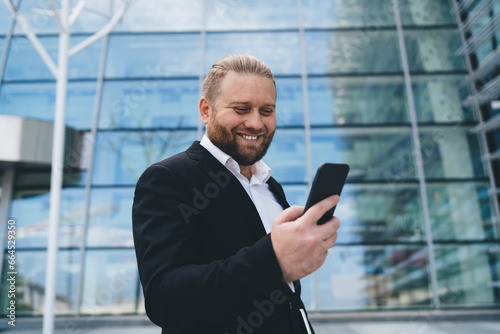Cheerful businessman standing and using smartphone opposite building