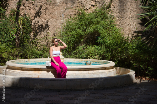 South American woman, young, beautiful, blonde with white top, pink pants and sunglasses, sitting in a nice fountain, sunbathing, relaxed and calm. Concept travel, destinations, Europe, vacation.