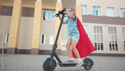 Girl superhero on electric scooter. happy family kid concept. a child on electric scooter rides on a large square in the city center under a blue sky with clouds and rays dream of sun. superhero girl