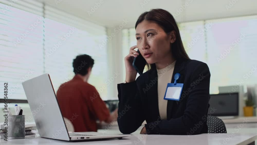 Medium shot of female office worker talking on smartphone while sitting at desk with wireless laptop Medium shot of female office worker talking on smartphone while sitting at desk with wireless laptop