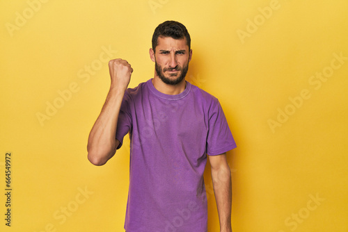 Young Hispanic man on yellow background showing fist to camera, aggressive facial expression.