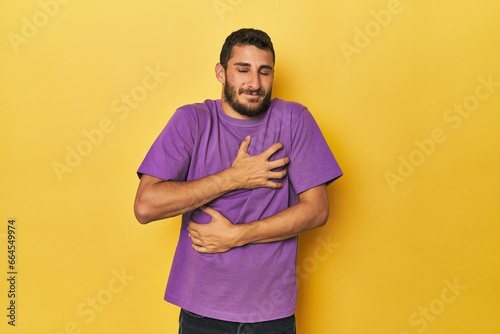 Young Hispanic man on yellow background laughs happily and has fun keeping hands on stomach.