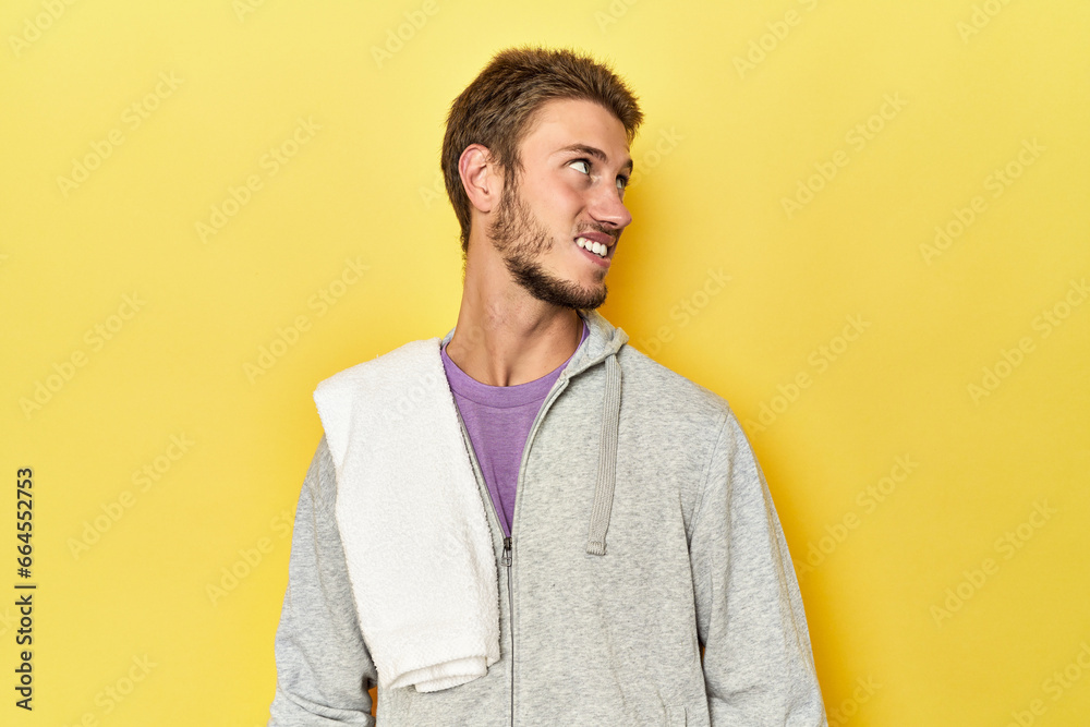 Sporty man in sweatshirt and towel on yellow looks aside smiling, cheerful and pleasant.