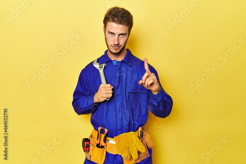 Technician in blue jumpsuit with tools on yellow showing number one with finger.