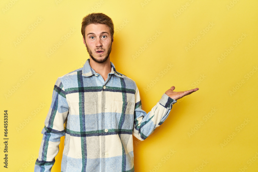 Young Caucasian man on a yellow studio background impressed holding copy space on palm. Young Caucasian man on a yellow studio background impressed holding copy space on palm.