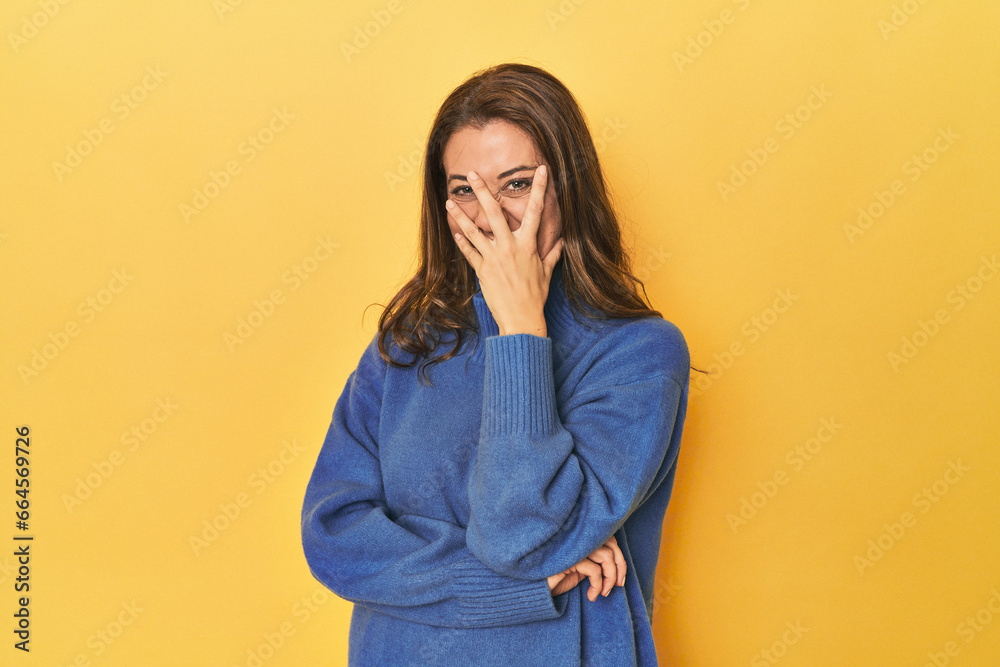 Middle-aged caucasian woman on yellow blink at the camera through fingers, embarrassed covering face.