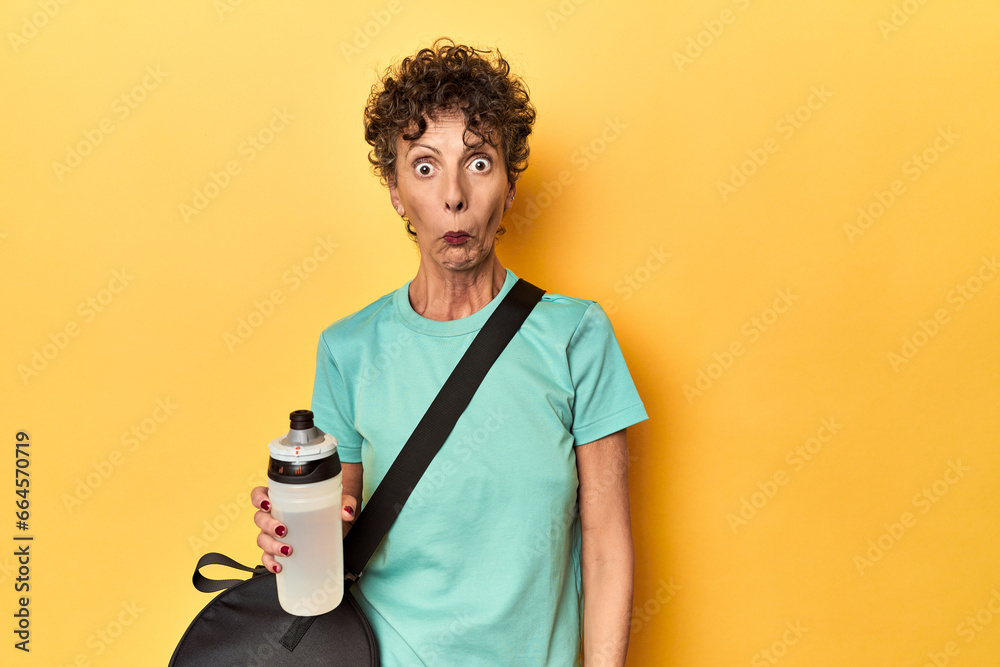 Sportswoman with gym bag & water on yellow studio shrugs shoulders and open eyes confused. Sportswoman with gym bag & water on yellow studio shrugs shoulders and open eyes confused.