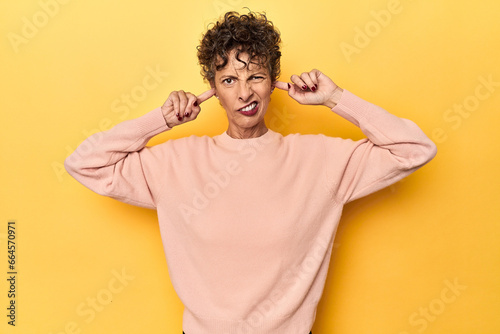 Mid-aged caucasian woman on vibrant yellow covering ears with fingers, stressed and desperate by a loudly ambient.
