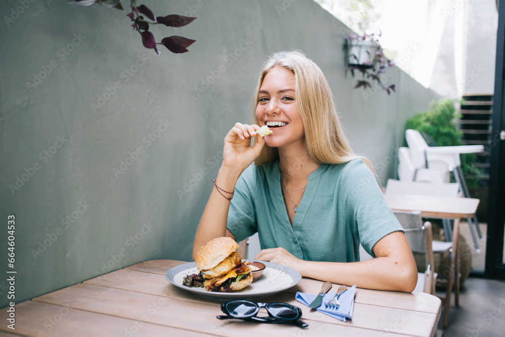 Portrait of cheerful female smiling at camera while eating appetizing burger with chicken posing at table with plate of unhealthy American dinner, happy hipster girl with hamburger have bite outdoors Portrait of cheerful female smiling at camera while eating appetizing burger with chicken posing at table with plate of unhealthy American dinner, happy hipster girl with hamburger have bite outdoors