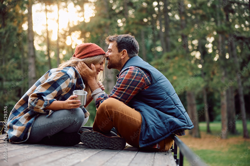 Affectionate man kissing his girlfriend in forehead while relaxing on top of their camper van in woods. Affectionate man kissing his girlfriend in forehead while relaxing on top of their camper van in woods.