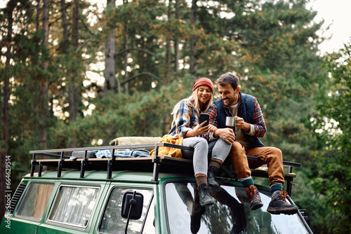 Happy couple using mobile phone while sitting on roof of their camper trailer in nature.
