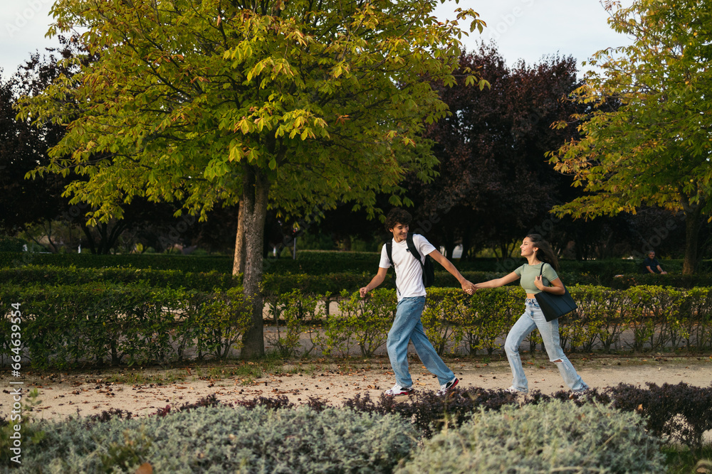 Fototapeta premium Pair of college students running together to university. They are smiling and holding hands while they run and hold their backpacks.