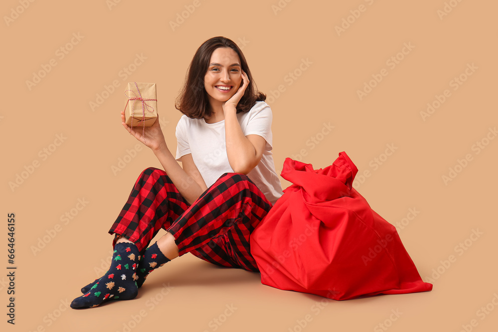 Beautiful young woman in pajamas with Santa Claus bag and gift box on brown background Beautiful young woman in pajamas with Santa Claus bag and gift box on brown background