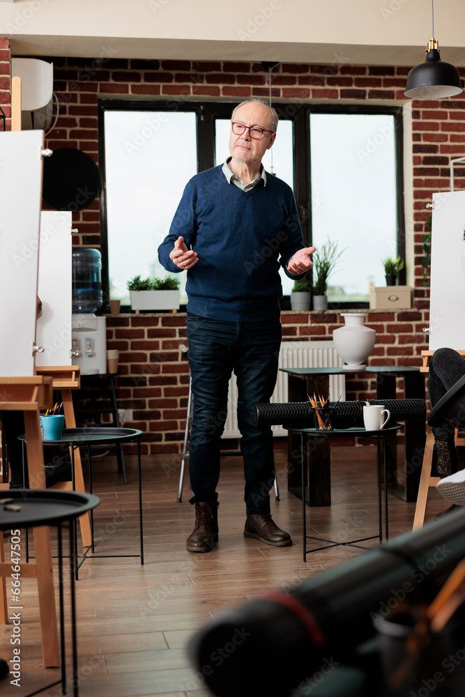 Senior man wearing glasses teaching art to adults in group class, standing in classroom introducing to drawing during creative workshop. Learning to draw from proffesional instructor Senior man wearing glasses teaching art to adults in group class, standing in classroom introducing to drawing during creative workshop. Learning to draw from proffesional instructor