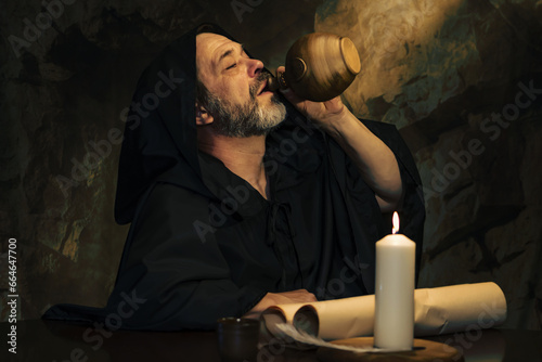 An elderly monk drinking from a clay jug, a dark and smoky stone cell, a candle burning.