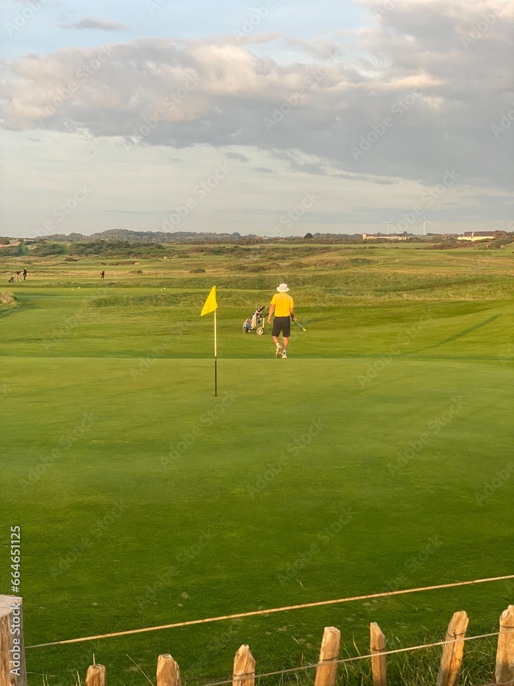 playing golf on a links course with a yellow top on in front of a yellow flag stood on the green hitting a putt with a putter. playing golf on a links course with a yellow top on in front of a yellow flag stood on the green hitting a putt with a putter.