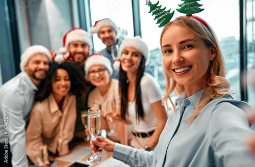 Successful cheerful happy fun business colleagues taking selfie together celebrating New Year's Eve. Beautiful blonde woman with decorative Christmas tree ears smiling and looking at the camera.