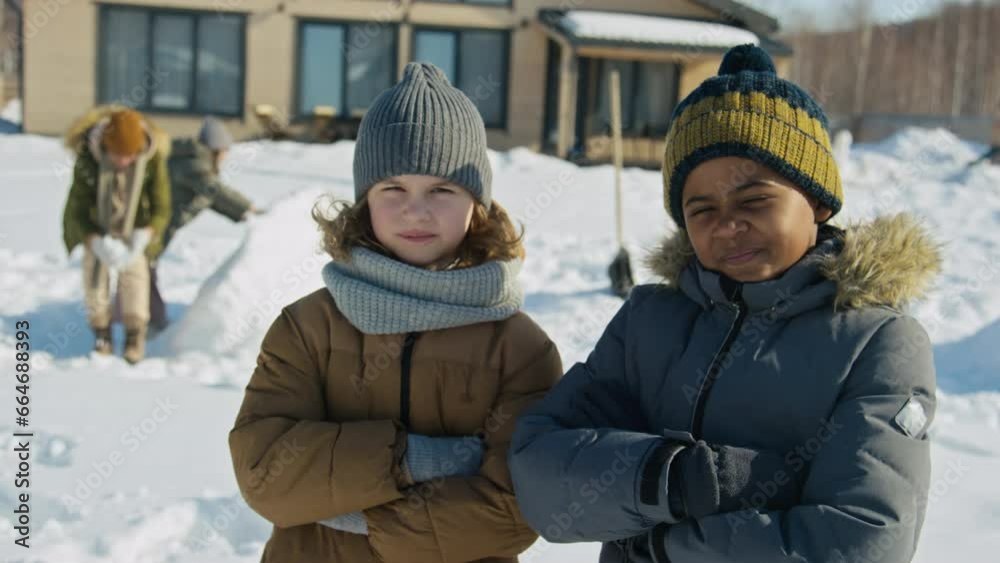 Medium portrait shot of two young mischievous Caucasian, African American boys in woolly hats and warm jackets standing in yard on sunny day, looking at camera, and girls building snow castle at back Medium portrait shot of two young mischievous Caucasian, African American boys in woolly hats and warm jackets standing in yard on sunny day, looking at camera, and girls building snow castle at back