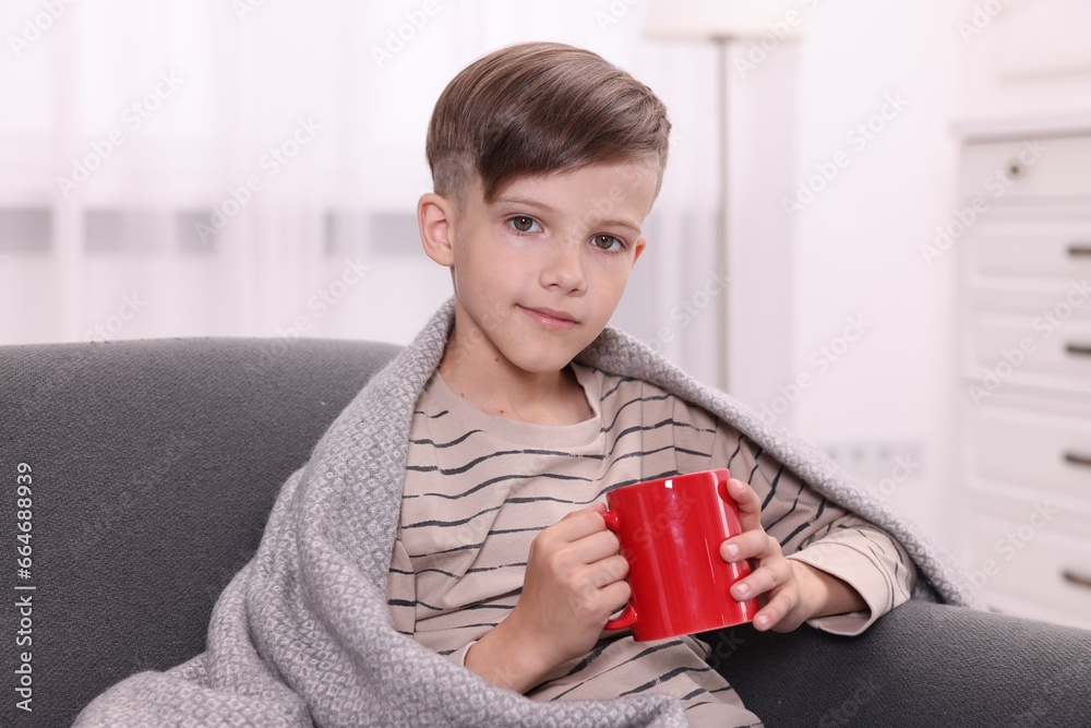 Cute boy under plaid with red ceramic mug on sofa at home