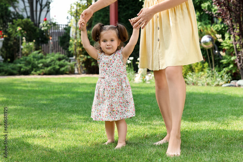 Mother supporting baby girl while she learning to walk on green grass in park