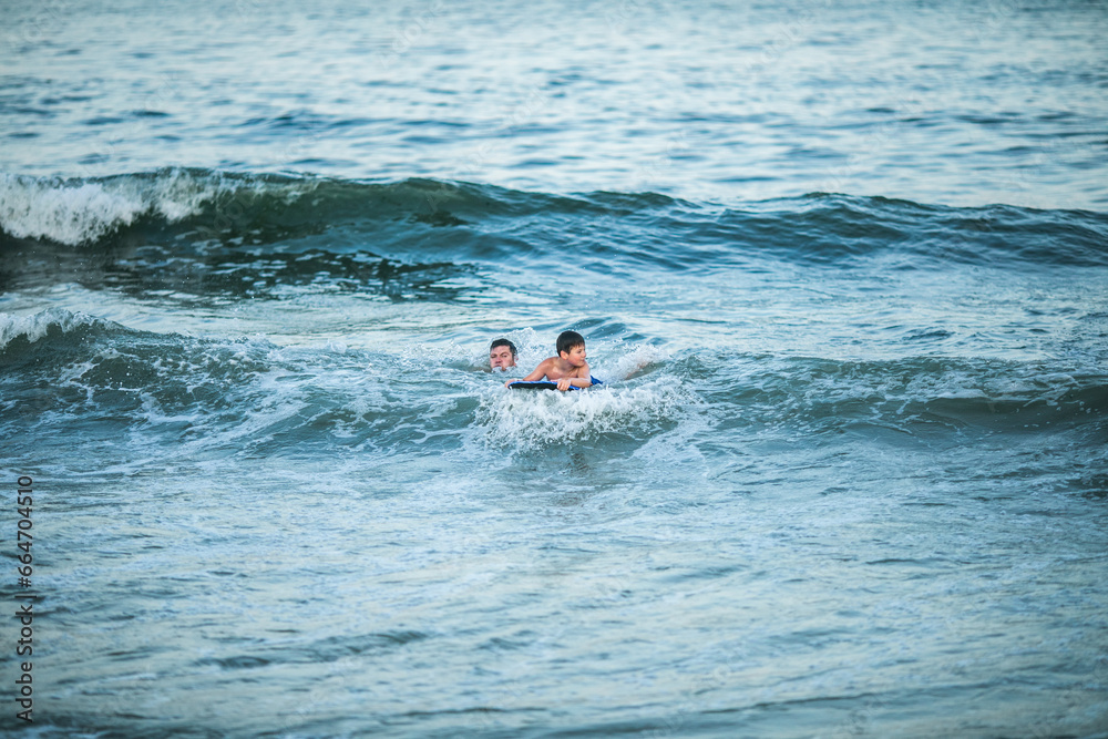Little surfer learn to ride on surfboard on sea wave. Father with son play in summer ocean, learning surfing. Little boy swim on surf board Little surfer learn to ride on surfboard on sea wave. Father with son play in summer ocean, learning surfing. Little boy swim on surf board