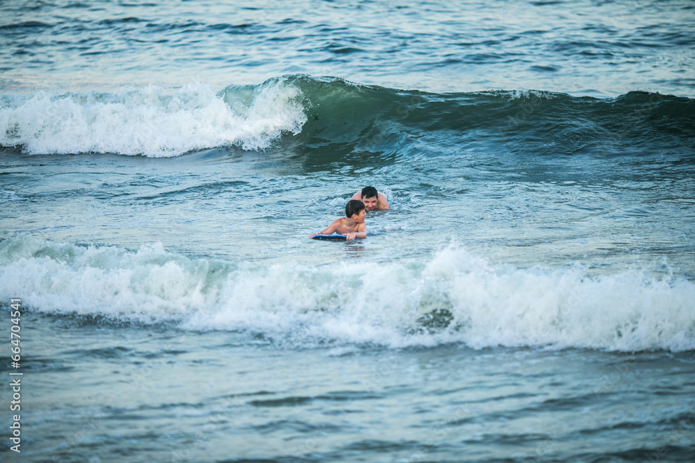 Little surfer learn to ride on surfboard on sea wave. Father with son play in summer ocean, learning surfing. Little boy swim on surf board Little surfer learn to ride on surfboard on sea wave. Father with son play in summer ocean, learning surfing. Little boy swim on surf board