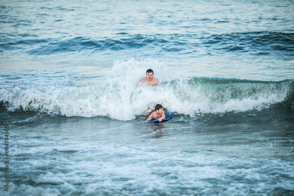 Little surfer learn to ride on surfboard on sea wave. Father with son play in summer ocean, learning surfing. Little boy swim on surf board Little surfer learn to ride on surfboard on sea wave. Father with son play in summer ocean, learning surfing. Little boy swim on surf board
