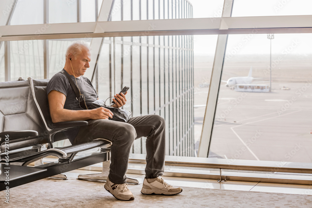 A serious gray-haired man wearing headphones looks at his smartphone, sitting at a panoramic wall in the airport departure hall