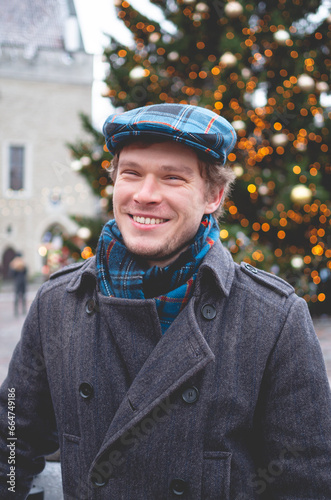 A handsome young man in a grey coat with a Scottish tweed cap and tartan scarf smiling while standing on a Tallinn Town Hall Square on a bright winter day with a Christmas tree in the background.
