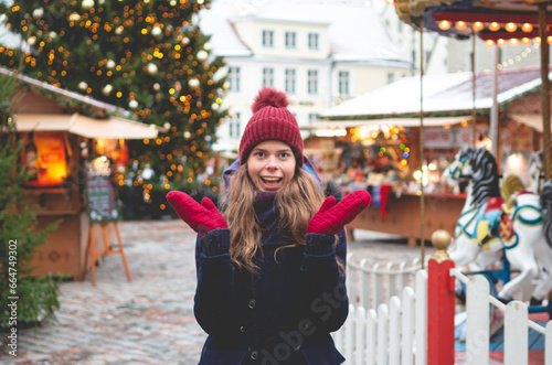 A surprised handsome young woman in a blue winter coat, red hat and gloves standing on a Tallinn Town Hall Square with a Christmas tree and a Christmas fare in the background