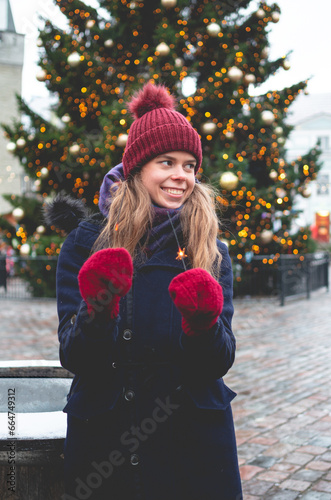 A young handsome smiling woman holding sparklers and standing on a Tallinn Town Hall Square on a cold winter day with a Christmas tree in the background