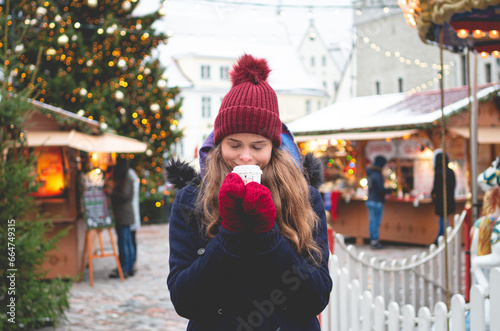 A handsome young woman in a blue winter coat, red hat and gloves enjoying cocoa and standing on a Tallinn Town Hall Square with a Christmas tree and a Christmas fare in the background