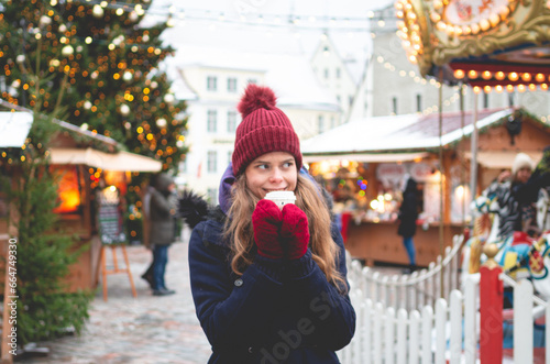 A handsome young woman in a blue winter coat, red hat and gloves holding coca cup and standing on a Tallinn Town Hall Square with a Christmas fare and a children's carousel in the background