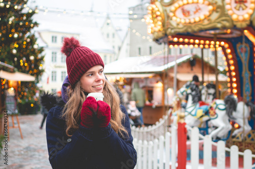 A handsome young woman in a blue winter coat, red hat and gloves holding coca cup and standing on a Tallinn Town Hall Square with a Christmas fare and a children's carousel in the background