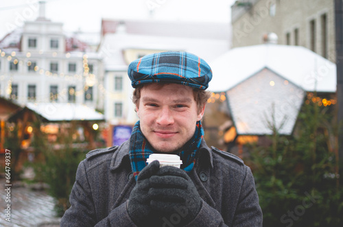 A handsome young man in a grey coat with a Scottish tweed cap and tartan scarf holding cocoa cup and standing on a Tallinn Town Hall Square on a winter day with a Christmas fare in the background