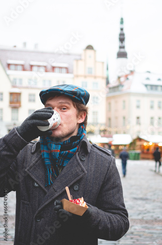 A handsome young man in a grey coat with a Scottish tweed cap and tartan scarf drinking cocoa and standing on a Tallinn Town Hall Square on a bright winter day with a Christmas fare in the background